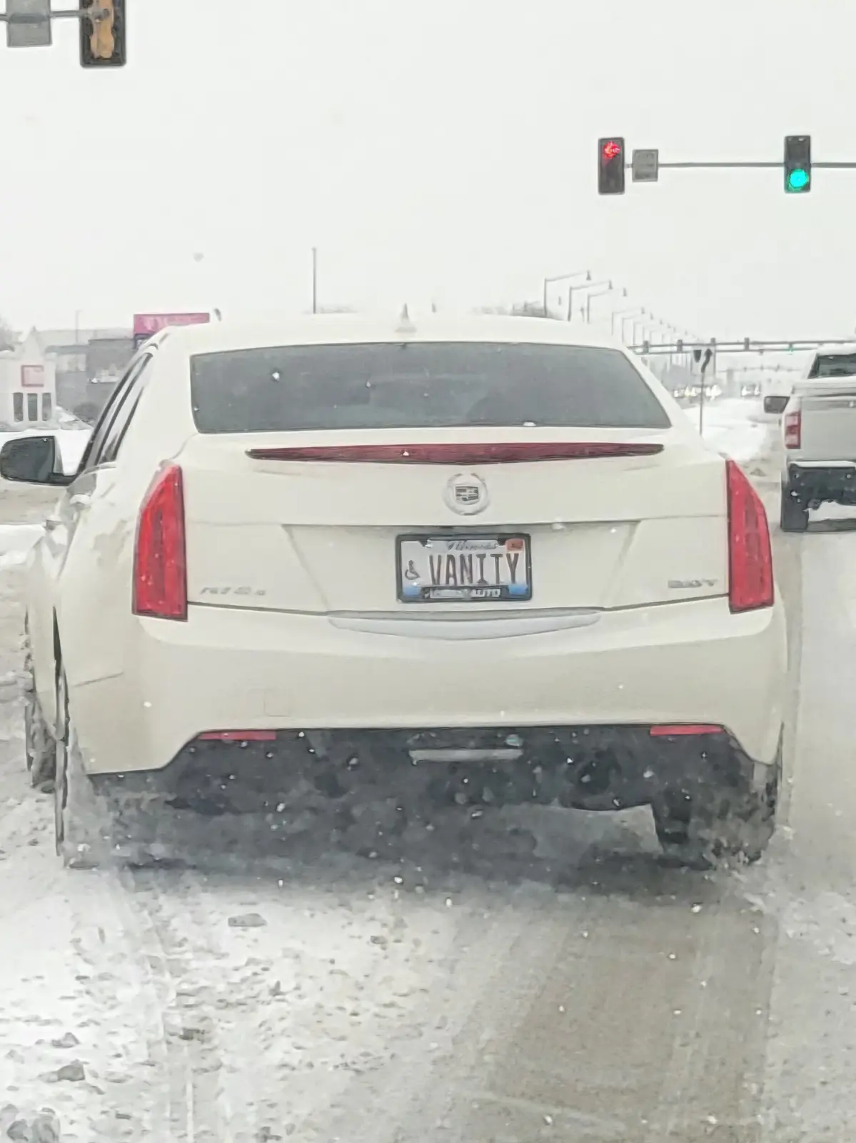 A white Cadillac sedan is stopped at a snowy intersection with traffic lights, its rear license plate reading 'VANITY' with…