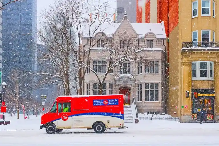A red and white canadian postal service truck drives through a snowy city street, passing a historic stone building and a…