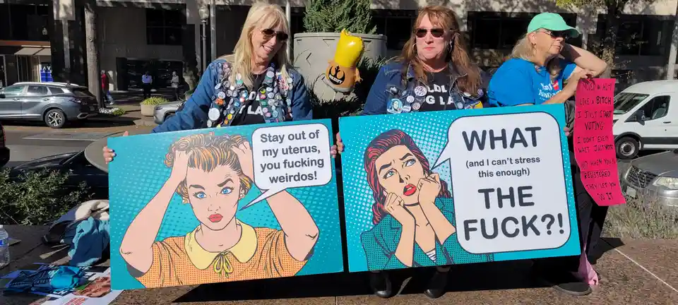 Three women at an outdoor protest hold large pop-art style signs featuring cartoon women with speech bubbles. The left sign…