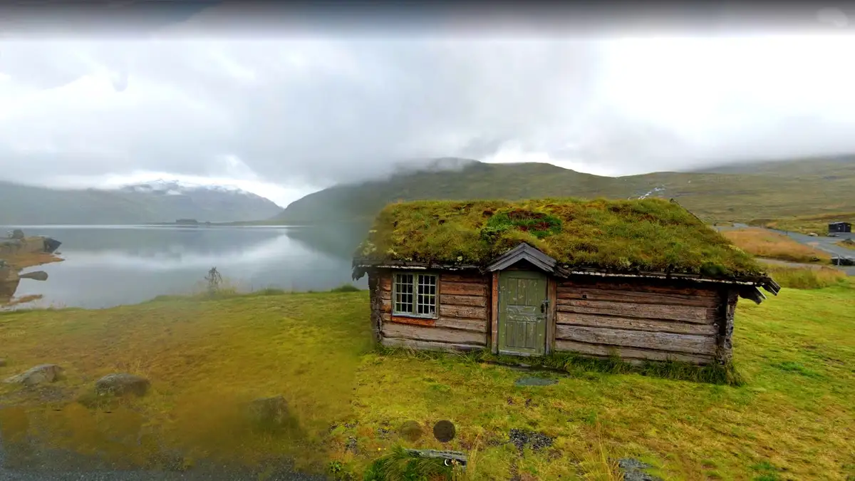 A rustic wooden cabin with a grassy roof sits beside a calm lake under a cloudy sky, with mountains in the background. The…