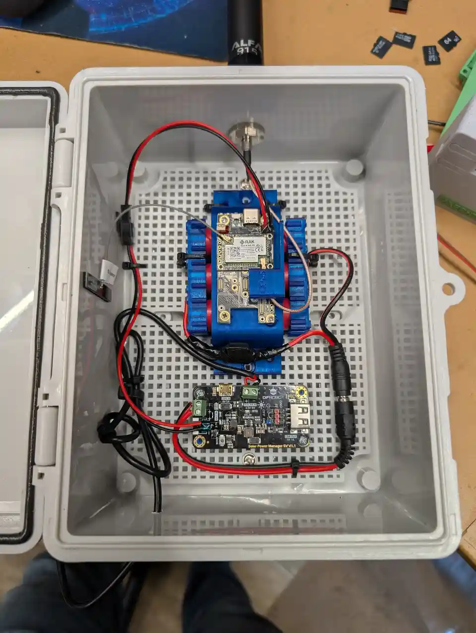 An overhead shot shows a small, blue 3D-printed robot with a circuit board and antenna inside a white plastic case. Wires…