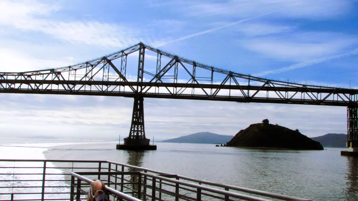 A scenic photo of a large steel truss bridge spanning a body of water under a partly cloudy sky, with a small island visible…