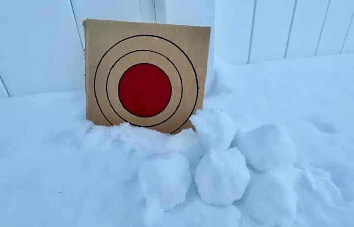 A cardboard target with concentric circles and a red center stands upright in deep snow, partially buried. Several lumpy…