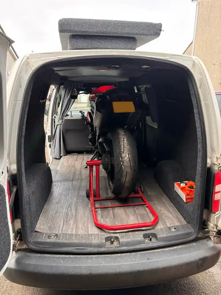 The rear of a white van is open, revealing a red motorcycle on a red stand inside. The van's interior has been modified with…