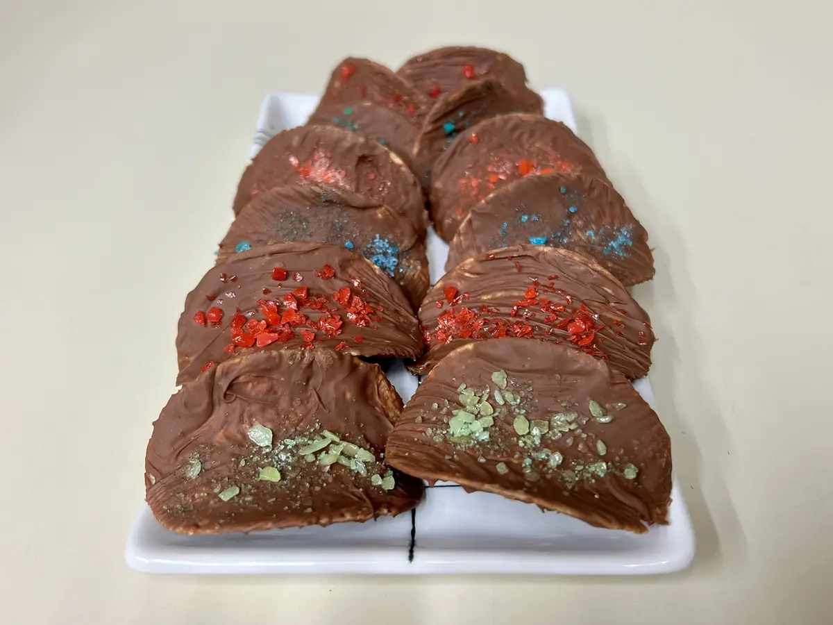 A white rectangular plate holds multiple chocolate-covered treats arranged in two rows. Each treat is topped with colorful…