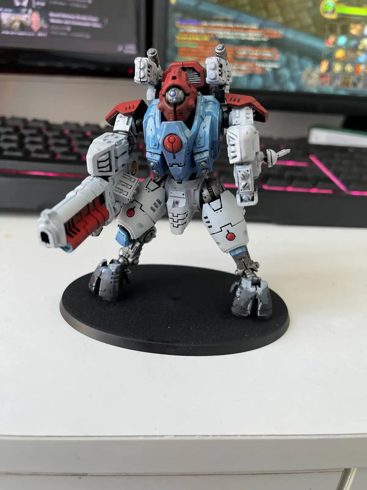 A painted, blue-and-red humanoid robot figurine stands on a black circular base on a white desk, with a keyboard and two…