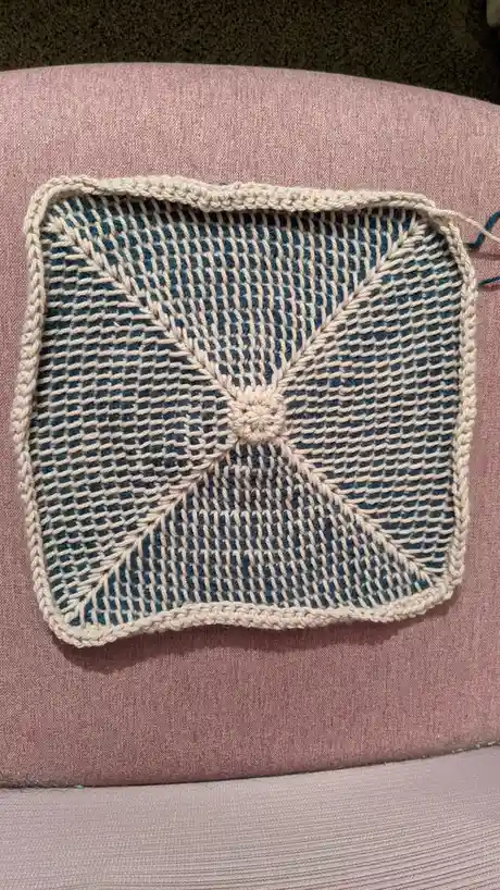 A square, hand-crocheted textile with a beige border and a central floral pattern made of beige and blue yarn, resting on a…