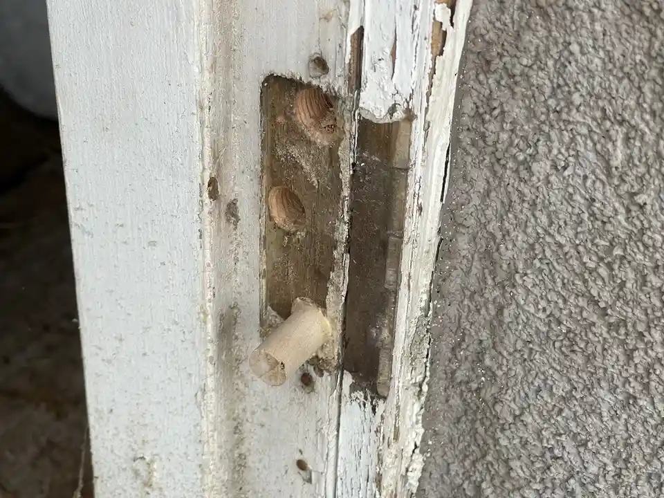 A close-up photo shows a weathered, white-painted wooden door frame with peeling paint and a damaged latch mechanism. The…
