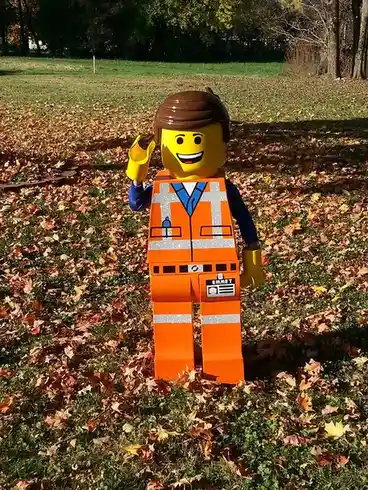 A life-sized cardboard cutout of the Lego character Emmet stands in a grassy field covered with fallen autumn leaves, waving…