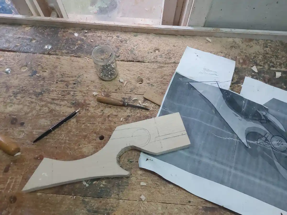 A woodworking bench shows a partially carved wooden piece shaped like a bat, next to a tracing paper with a detailed bat…