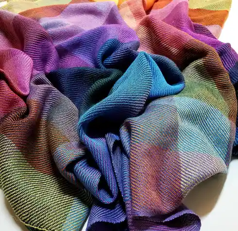 A close-up photo shows a pile of colorful, textured fabric scarves in a rainbow gradient, from yellow to purple to blue…