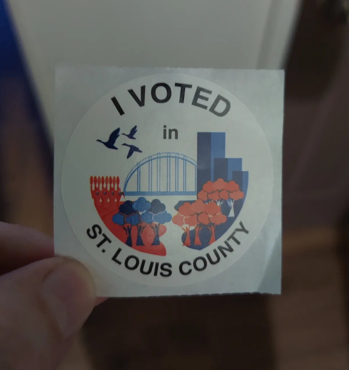 A close-up photo shows a hand holding a circular white sticker. The sticker reads 'I VOTED in ST. LOUIS COUNTY' in bold…
