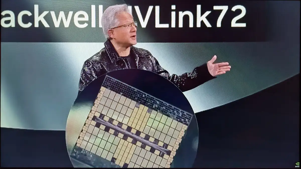 A man with gray hair and glasses, wearing a shiny black jacket, stands on a stage gesturing toward a large circular wafer…
