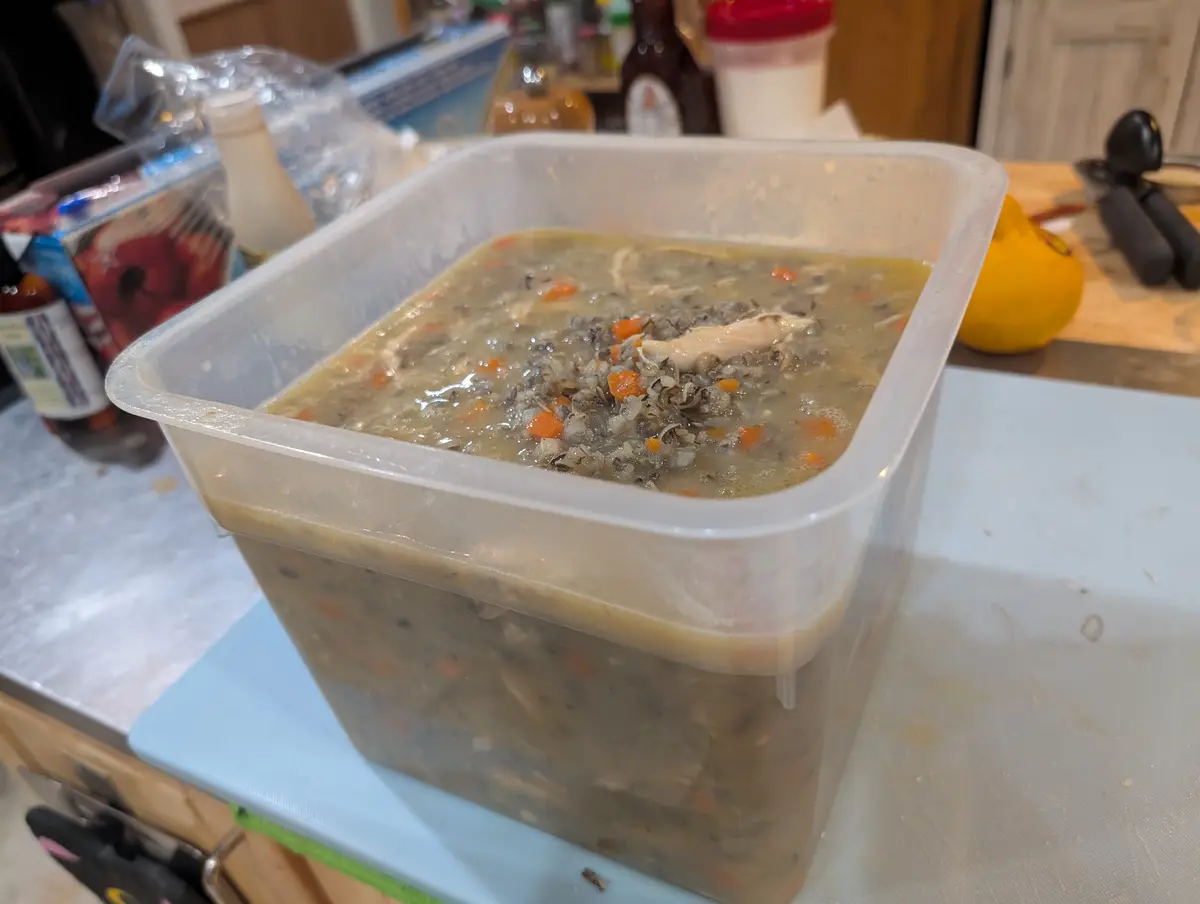 A large, clear plastic container filled with a thick, chunky soup containing visible pieces of chicken, carrots, and wild…