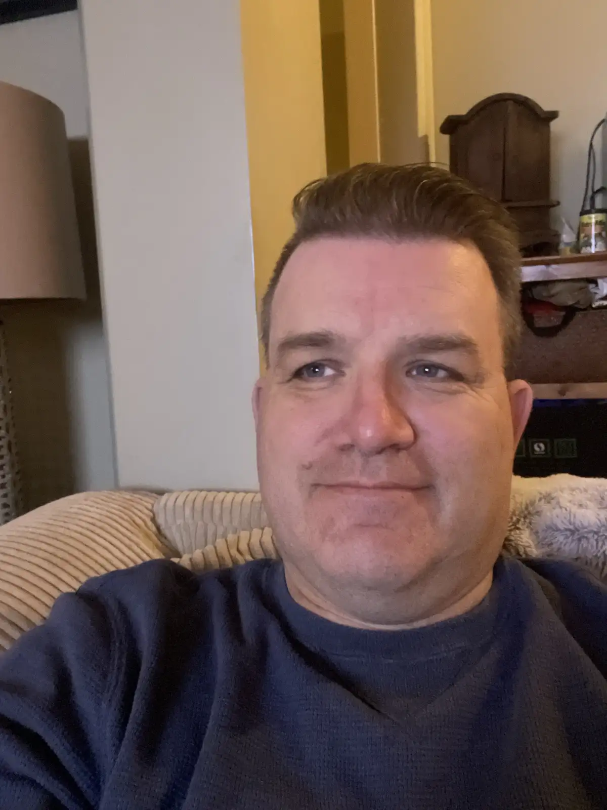 A man with short brown hair and a slight smile sits on a beige corduroy couch, wearing a dark blue sweatshirt. Behind him, a…