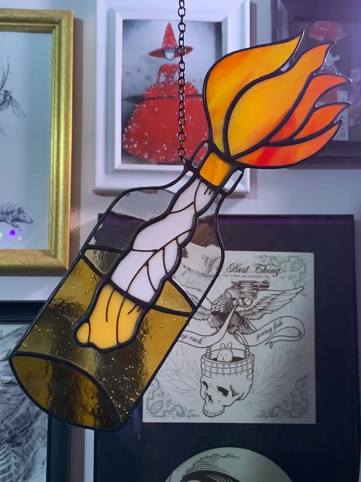 A stained glass suncatcher shaped like a bottle with a flaming top, hanging by a chain against a wall with framed artwork…