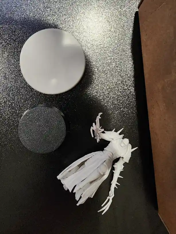 A white, unpainted fantasy miniature figure with spiky armor and flowing cape lies on a dark, textured surface next to two…