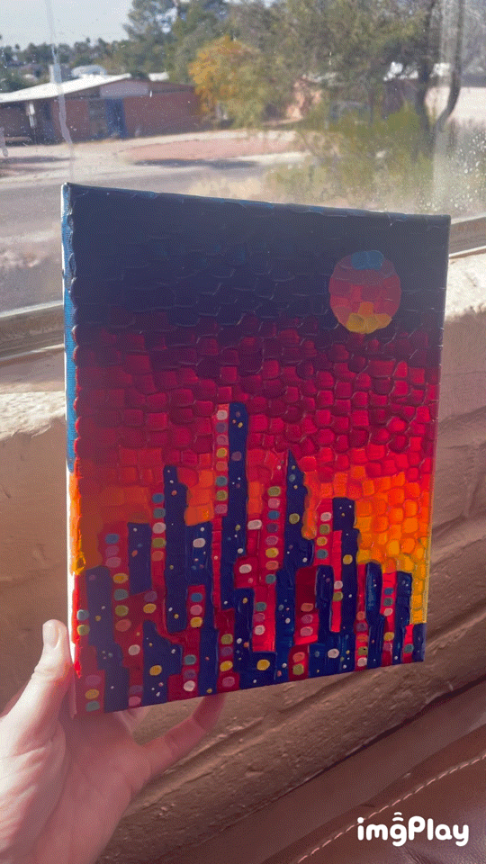 A hand holds a textured, colorful painting of a city skyline at sunset, with the sun depicted as a multi-colored circle. The…