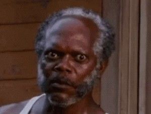 A close-up of a Black man with gray hair and a beard, wearing a white tank top, staring with wide, shocked eyes. The…