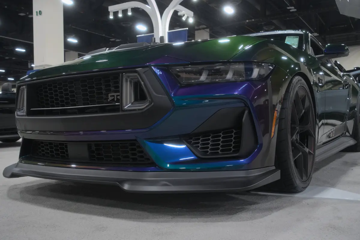 a close-up of a shiny, iridescent green and purple Ford Mustang Shelby GT500 on display at an auto show, with overlaid text…