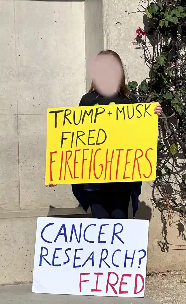 A person with a blurred face holds a yellow sign reading 'TRUMP + MUSK FIRED FIREFIGHTERS' while a second white sign on the…