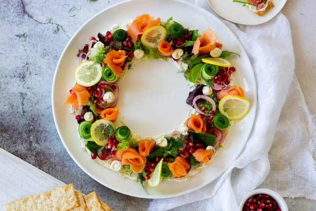 a circular platter of food arranged in a wreath shape with salmon, lemon slices, pomegranate seeds, and greens, leaving a…