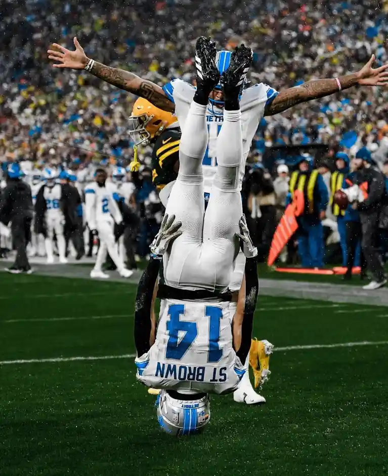 A football player wearing a white jersey with the number 14 and name 'ST BROWN' on the back is being held upside down by…
