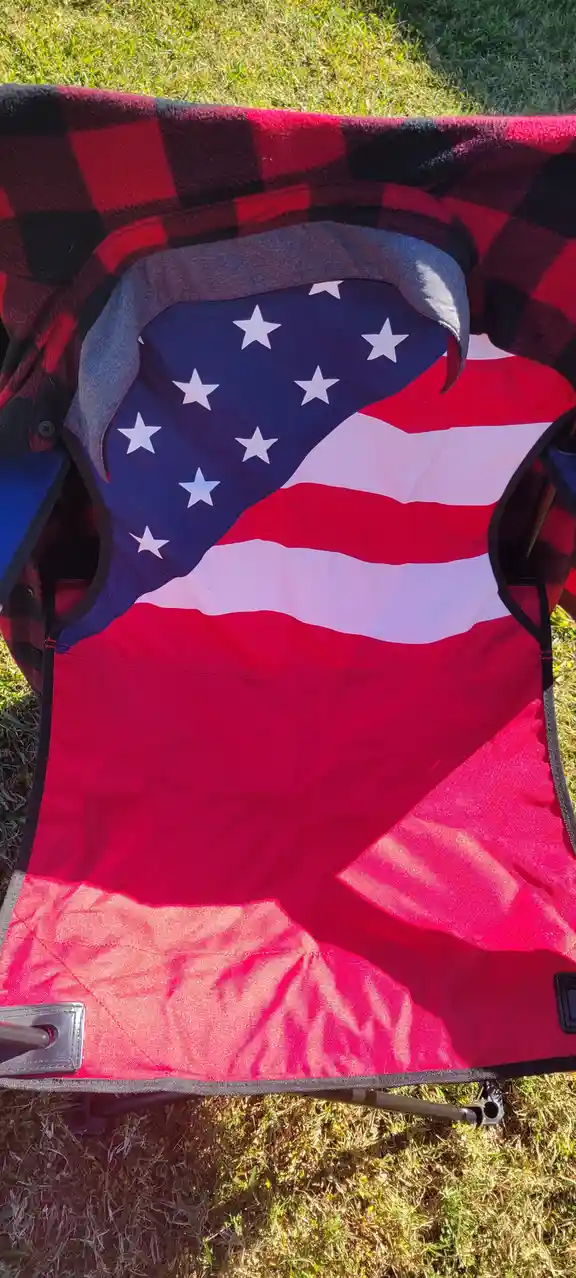 A red and black plaid blanket is draped over a folding chair outdoors on grass, partially covering an American flag that is…