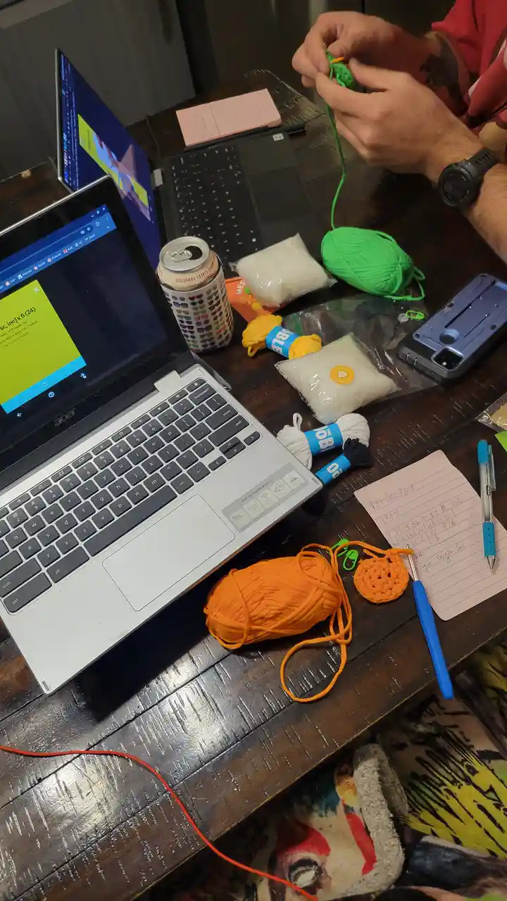 A person is crocheting with green yarn at a cluttered wooden table, surrounded by multiple laptops, skeins of yarn in…