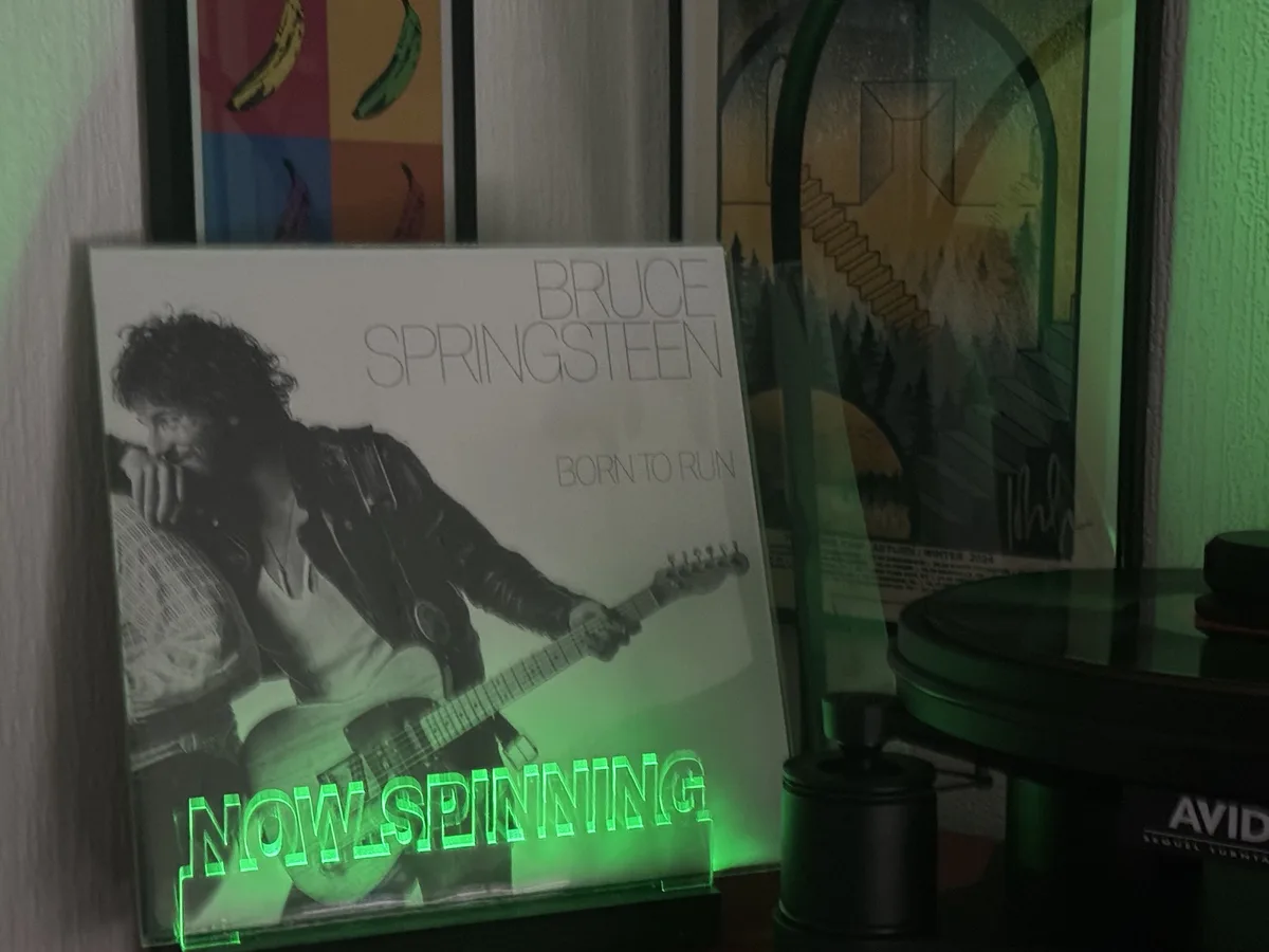 A black-and-white album cover of Bruce Springsteen's 'Born to Run' is displayed upright, with a glowing green neon sign…