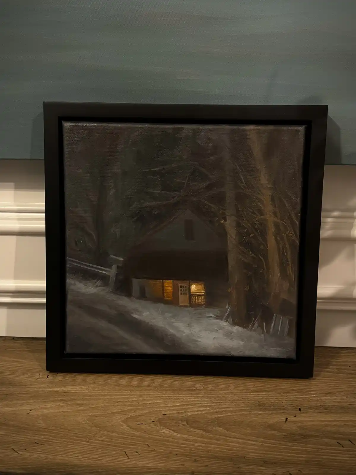 A framed painting depicts a cozy house with warm lights glowing from its windows, set against a snowy, dark forest at night…