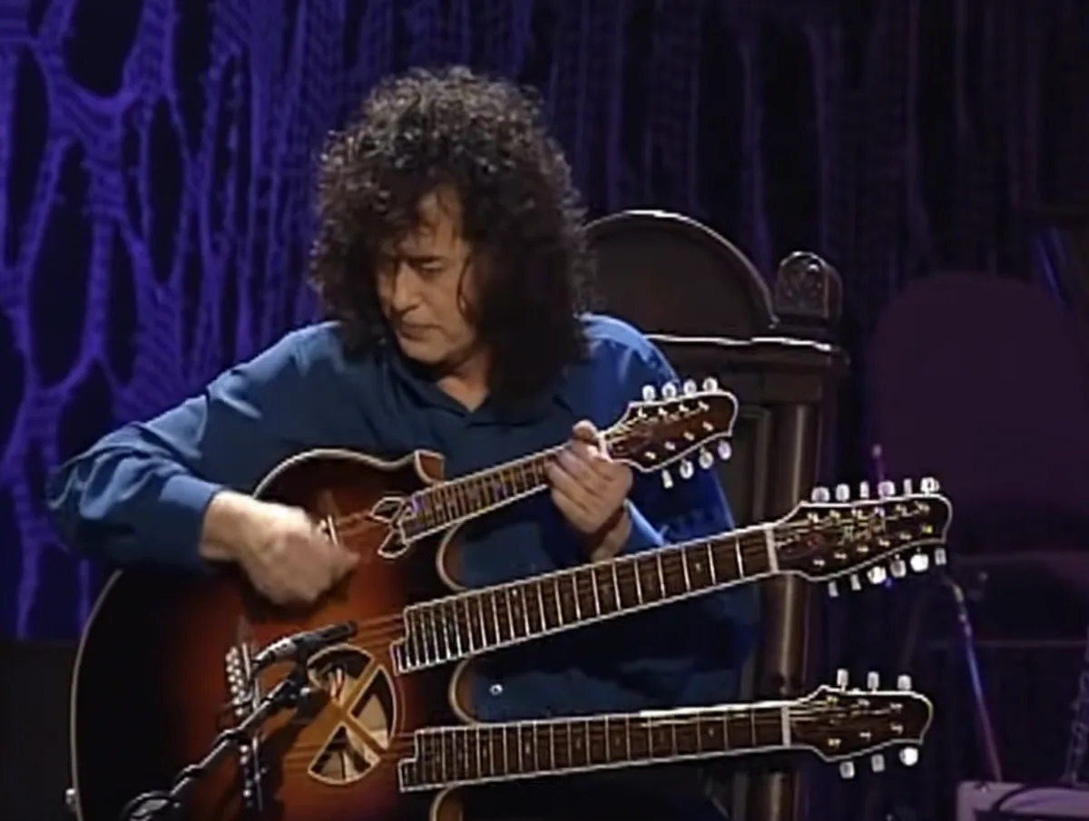 a man with curly dark hair wearing a blue shirt sits in a chair on a stage with a purple patterned backdrop, playing a…