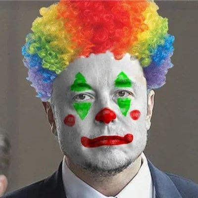A man in a suit and white face paint with green triangles, red nose, red lips, and red cheeks wears a rainbow-colored curly…