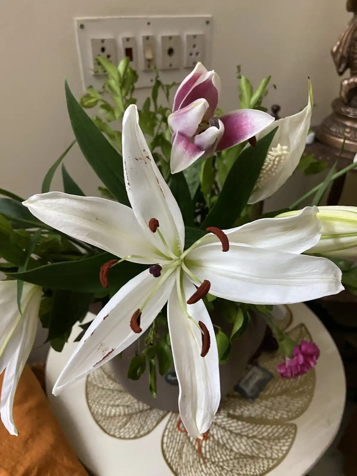 A close-up photo of a bouquet of lilies in a decorative vase, featuring a prominent white lily with brown speckles and…