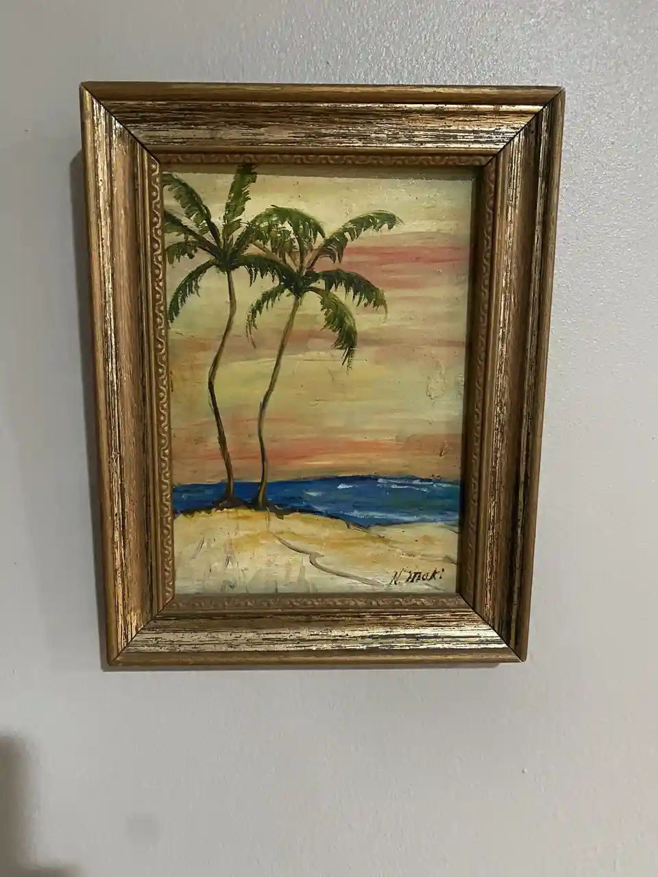 A framed painting of a tropical beach scene with two leaning palm trees on a sandy shore, a blue ocean, and a sunset sky…