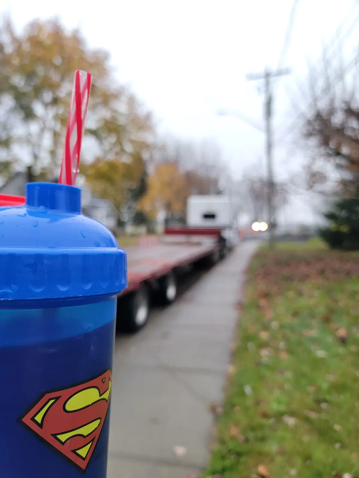 A close-up of a blue Superman-themed sippy cup with a red and white striped straw, positioned in the foreground. The…