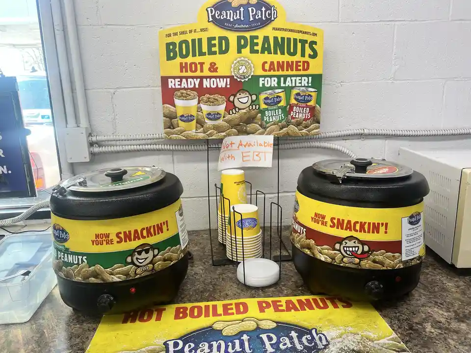 Two large black Peanut Patch hot peanut dispensers sit on a counter with a sign above them advertising boiled peanuts. A…