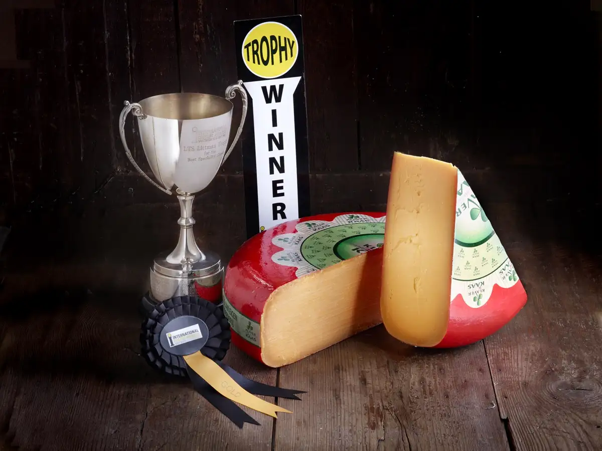 A large, round wheel of cheese with a wedge cut out sits next to a silver trophy cup and a black-and-white 'TROPHY WINNER'…