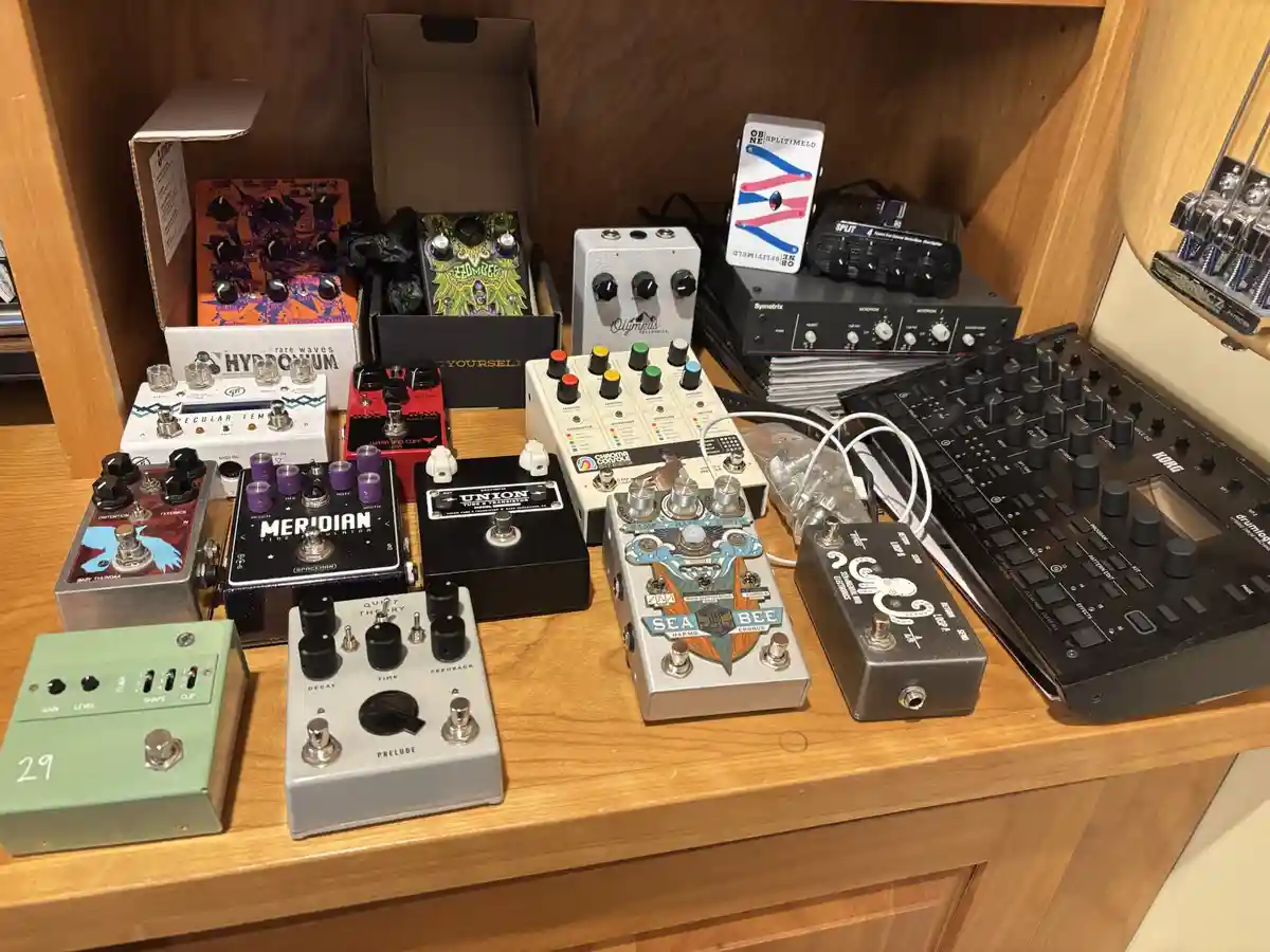 A wooden shelf holds a large collection of colorful guitar effect pedals and related gear, including a Korg synthesizer and…