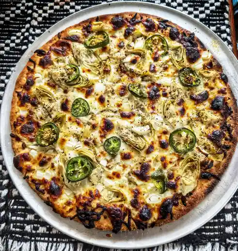 An overhead shot of a round pizza on a metal tray, featuring melted cheese, artichoke hearts, and sliced jalapeños, with…