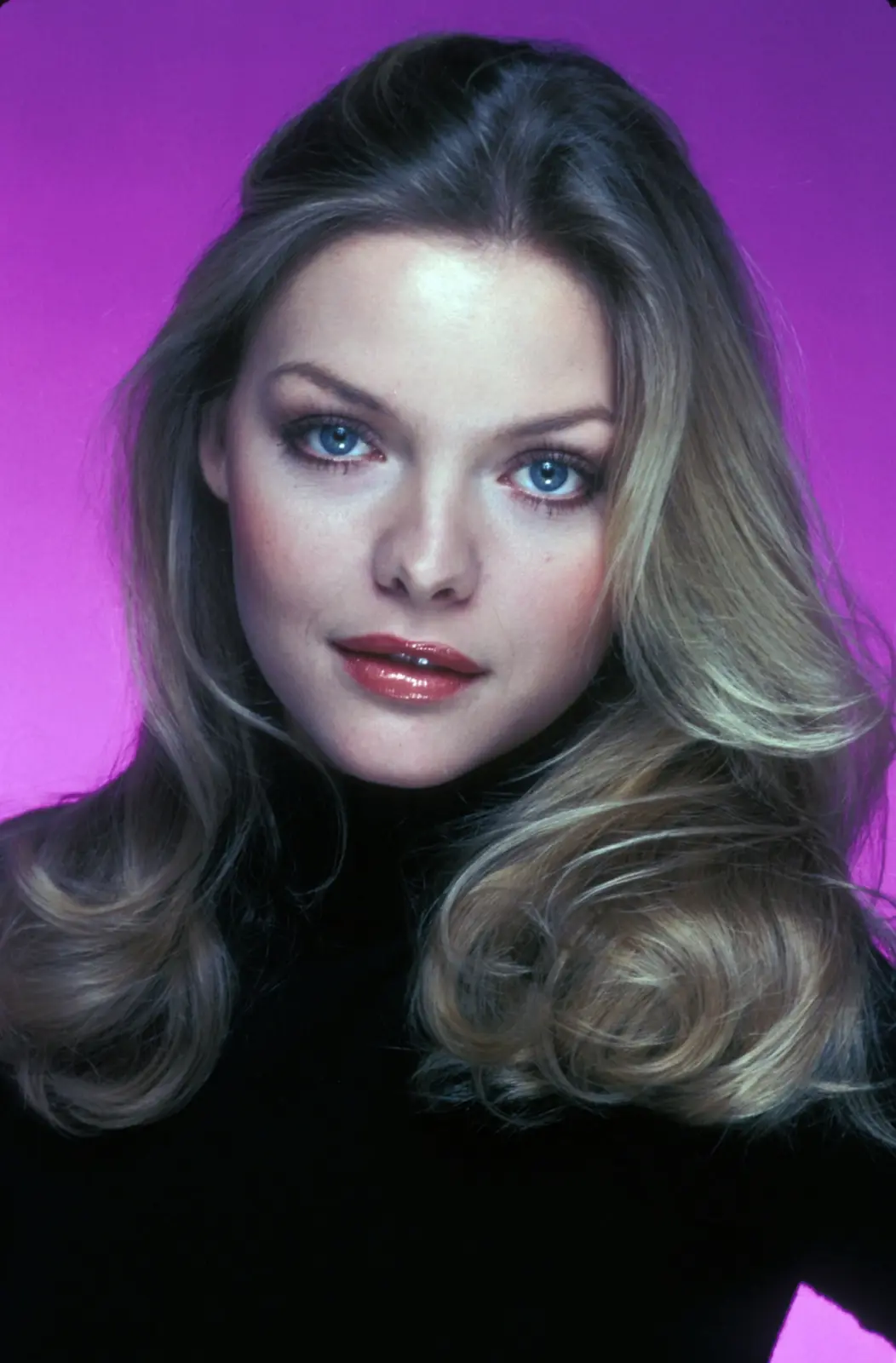 A vintage portrait of a woman with blonde hair and blue eyes against a vibrant purple background, wearing a black top. The…