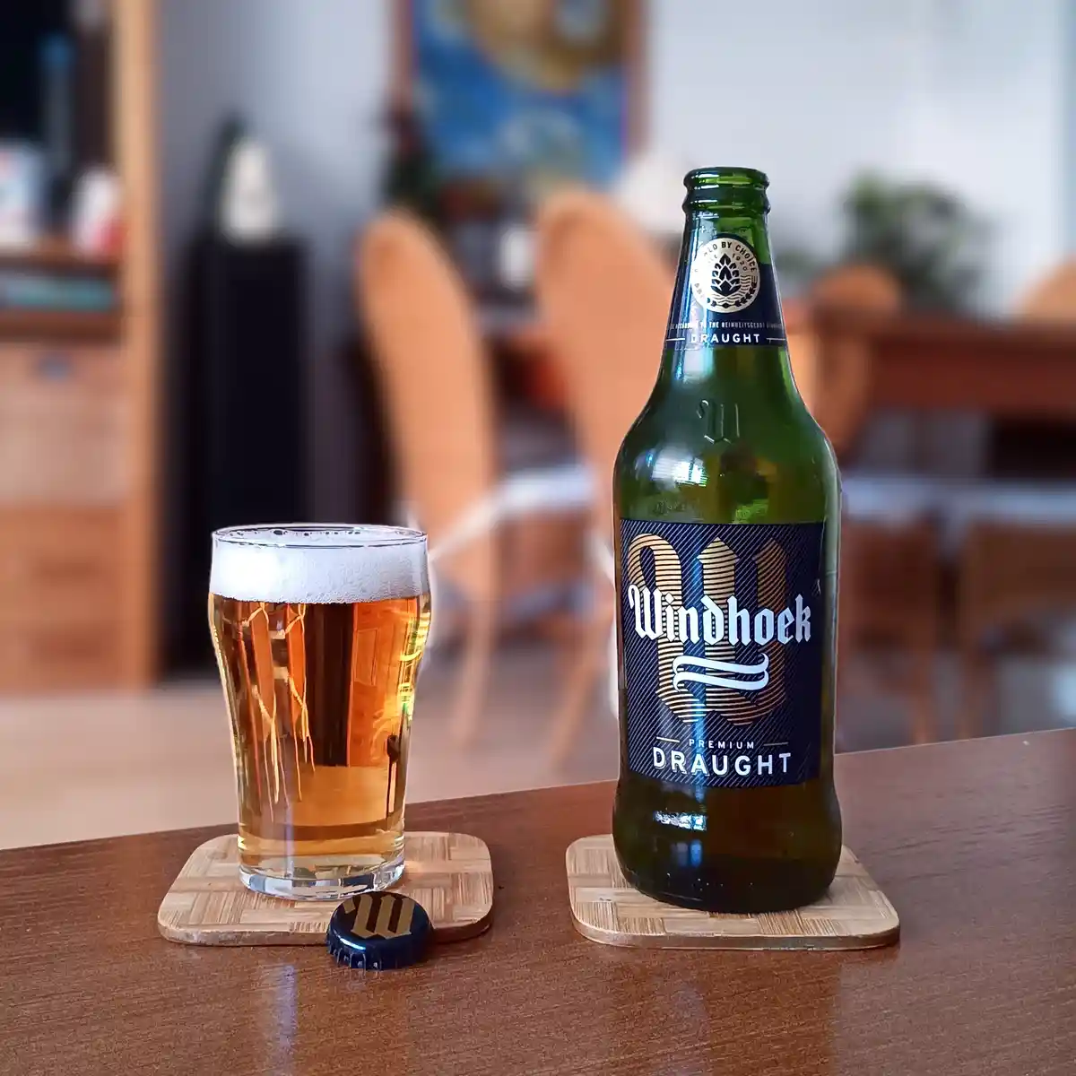 A green bottle of Windhoek Premium Draught beer and a full glass of beer sit on wooden coasters on a wooden table, with the…