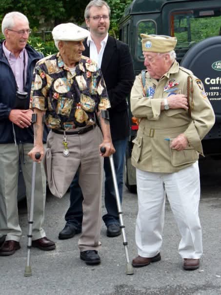 Four men stand outdoors near a Land Rover; two elderly veterans in decorated uniforms and using canes are central, flanked…