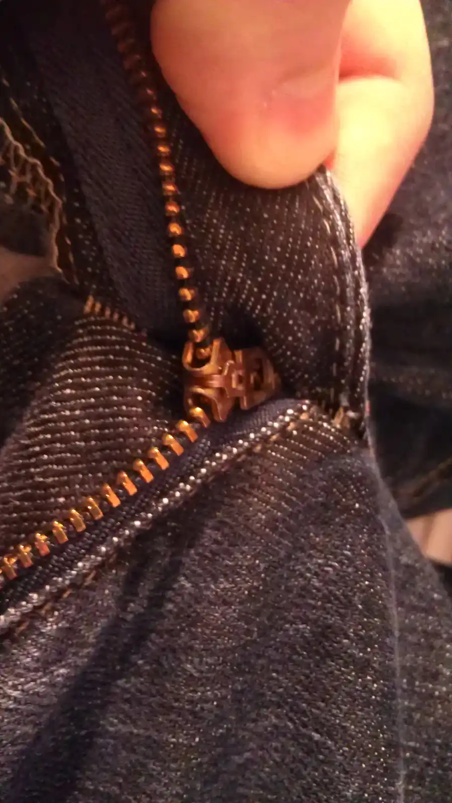 A close-up photo shows a person's thumb pressing down on the zipper pull of dark denim jeans, with the zipper partially…