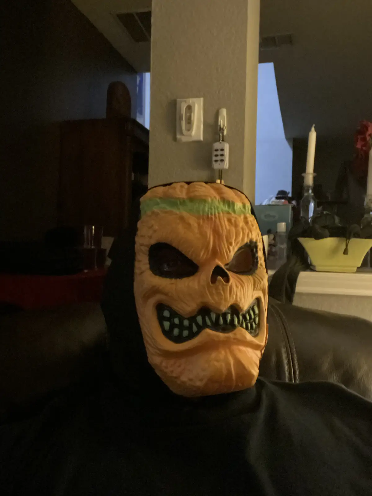 A person wearing a scary, orange pumpkin mask with green teeth and a green band across the forehead sits on a couch in a…
