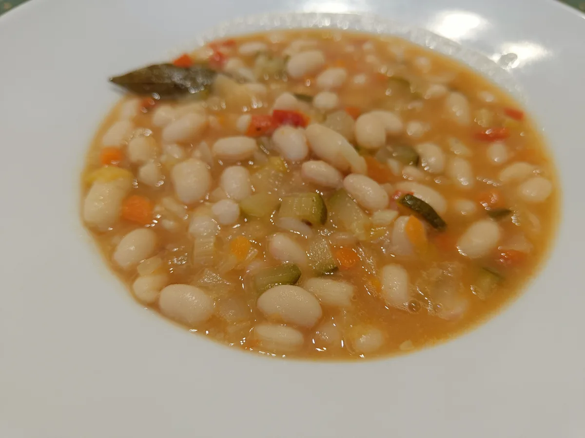 a close-up photo shows a white bowl filled with a light orange broth containing numerous white beans, small diced carrots…