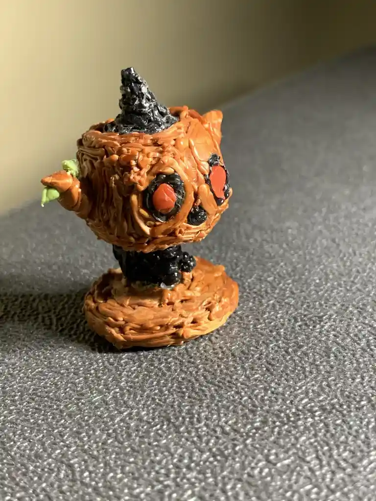 A small, hand-painted figurine resembling a pumpkin with a black witch's hat, red eyes, and a textured orange body stands on…