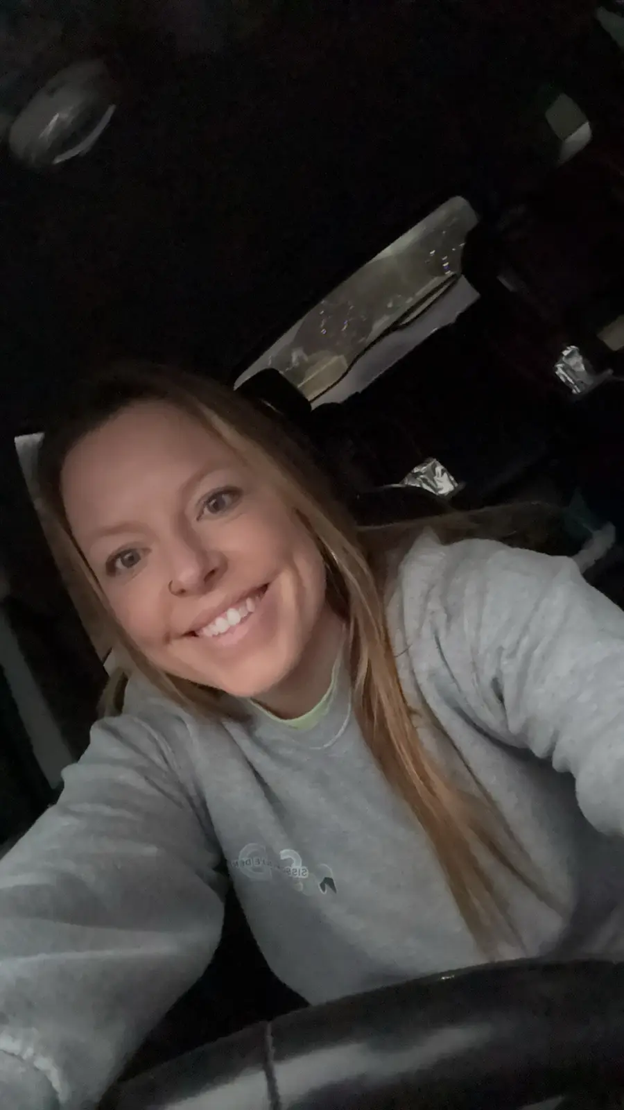 A selfie of a smiling woman with blonde hair wearing a grey sweatshirt, taken inside a car at night. The car's interior is…