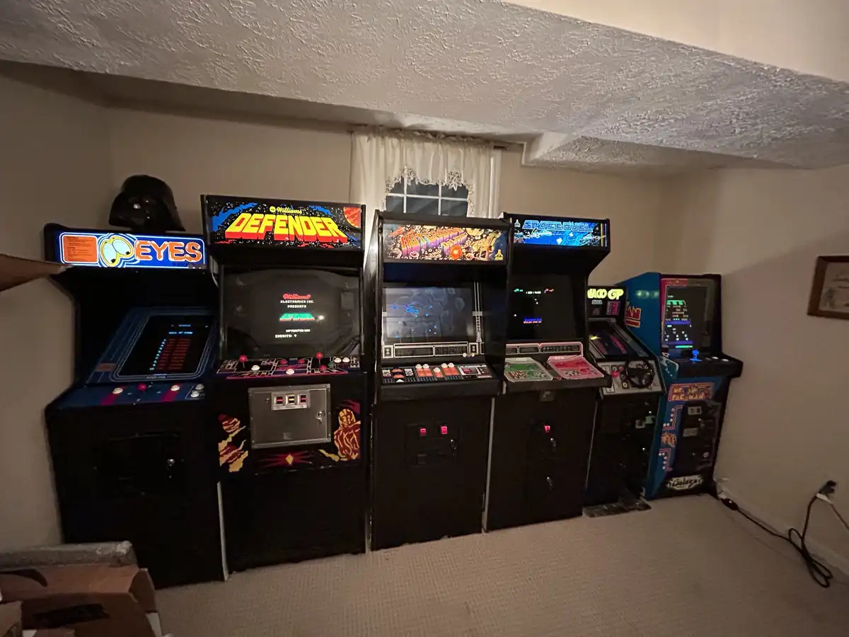 A row of vintage arcade machines, including Defender, Asteroids, and Space Duel, are lined up in a basement room. A hand…