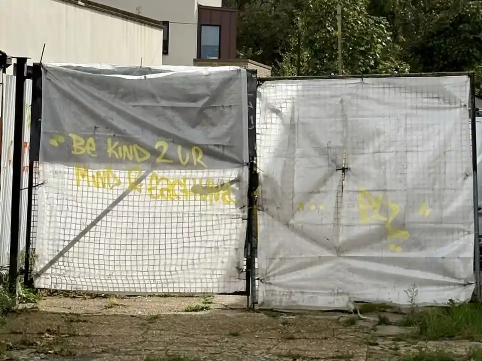 Two weathered white tarps hang on a fence, one with yellow spray-painted text reading 'Be kind 2 ur MIND & EARTH' and the…